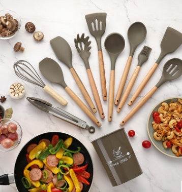Silicone kitchenware set with wooden handles displayed on a countertop with various ingredients, featuring utensils like spatulas, spoons, and a whisk.