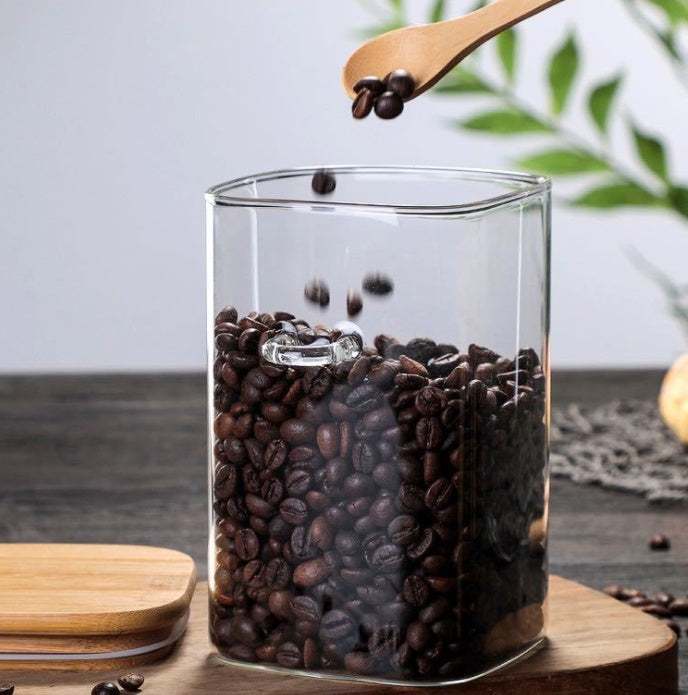 Glass square kitchen food storage can filled with coffee beans, wooden lid on side.