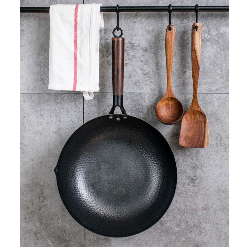 Traditional iron wok with wooden handle, hanging on a kitchen wall with utensils; high-quality, chemical-free cookware.