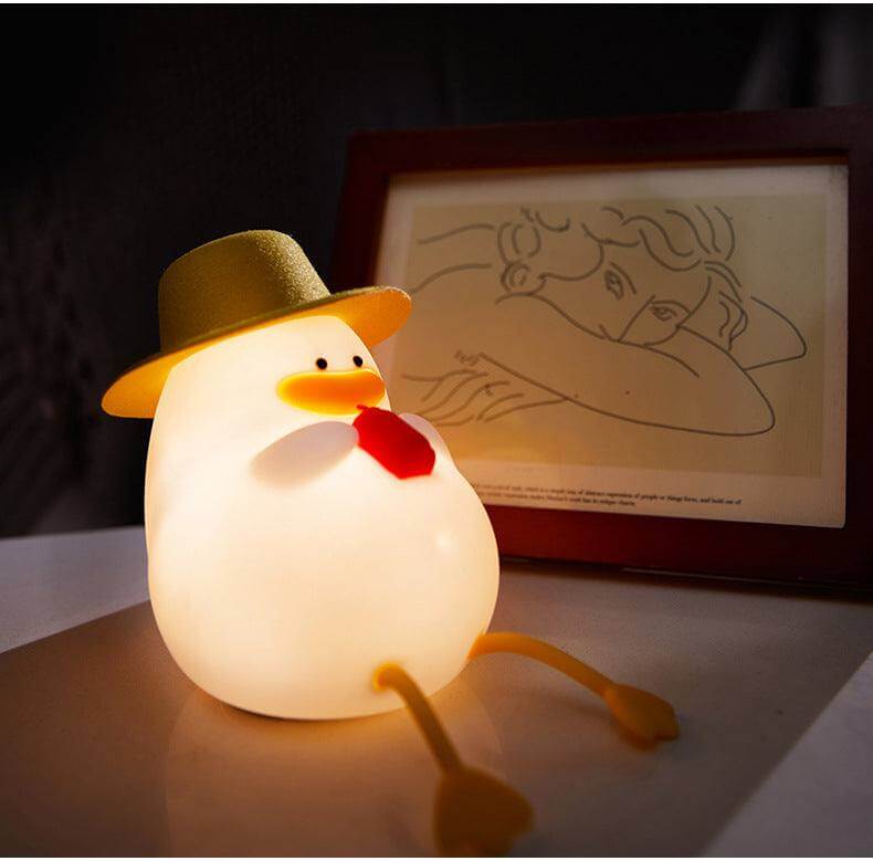 Duck-shaped silicone night lamp with a hat and tie, placed on a table next to a framed artwork. 