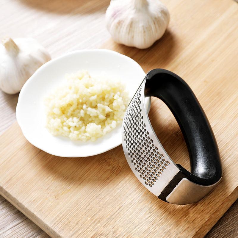 Stainless steel garlic press with ergonomic handle on a kitchen board, next to a plate of minced garlic.