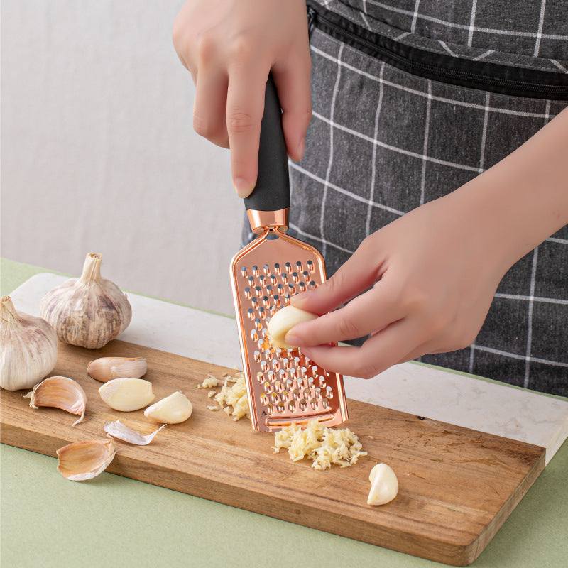 Copper-plated kitchen peeler grating garlic on wooden board.