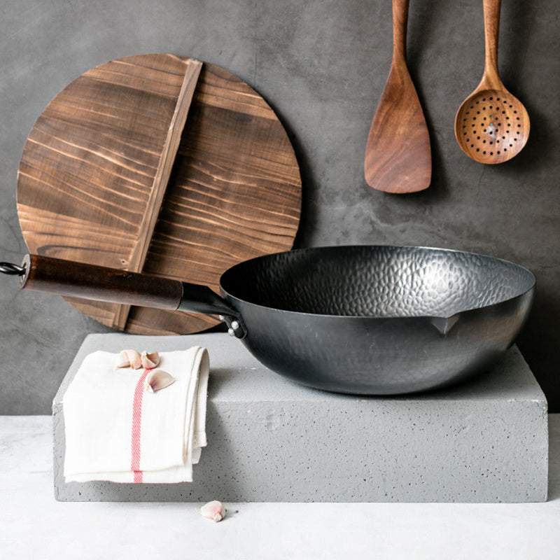 Iron pan with wooden handle and lid, displayed on a concrete block with hanging wooden utensils.