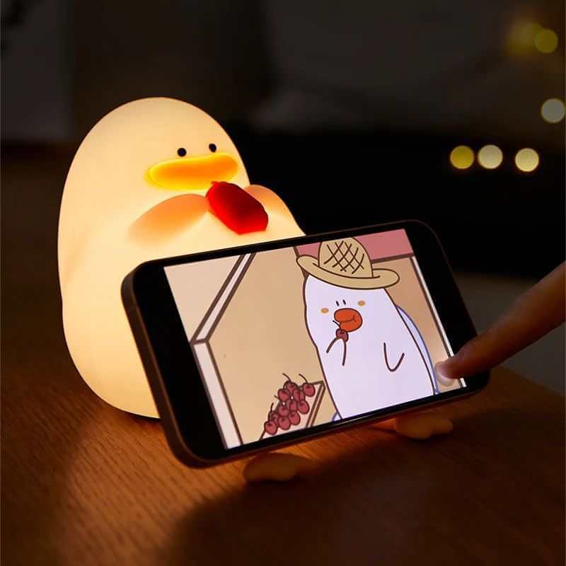 Duck-shaped silicone night lamp holding a mobile phone, illuminating softly on a table. 