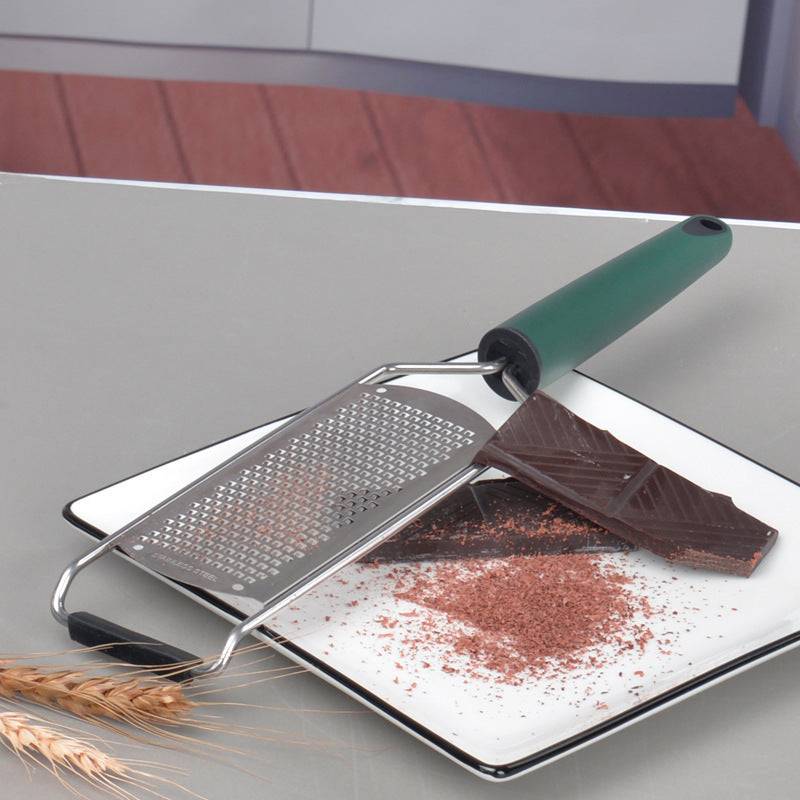 Stainless steel cheese grater with green handle, shown grating chocolate on a kitchen countertop.